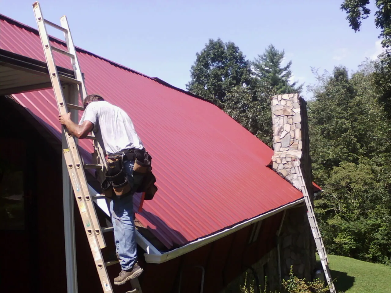 Roofer on a ladder beside a red metal roof and stone chimney — Soffit & Fascia Repair by Roofing Brothers in Leitchfield